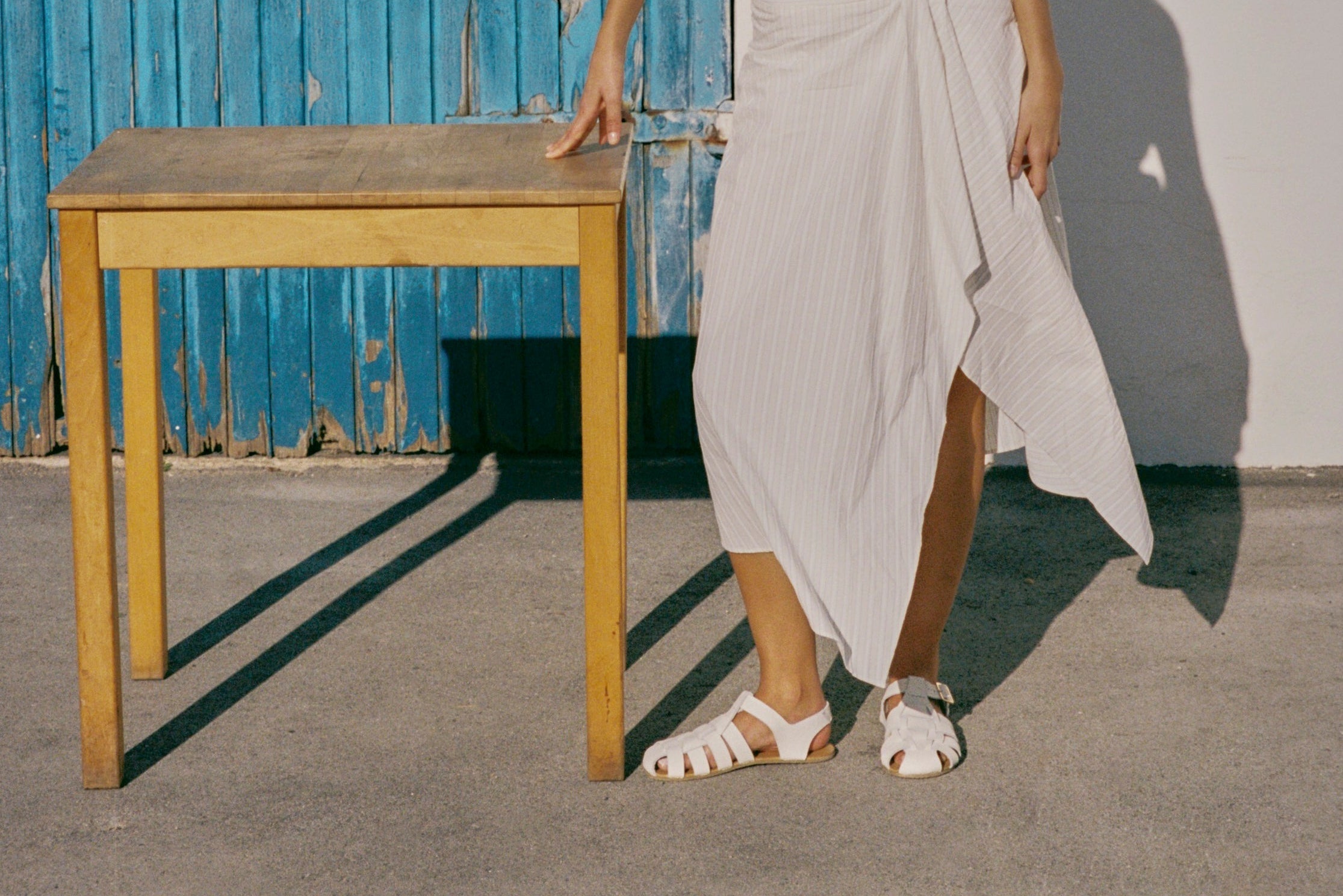 A woman stands outdoors next to a wooden table, wearing a white sleeveless ribbed knit top and a matching white wrap-style midi skirt with white sandals. The background features a blue corrugated metal door and a white wall.