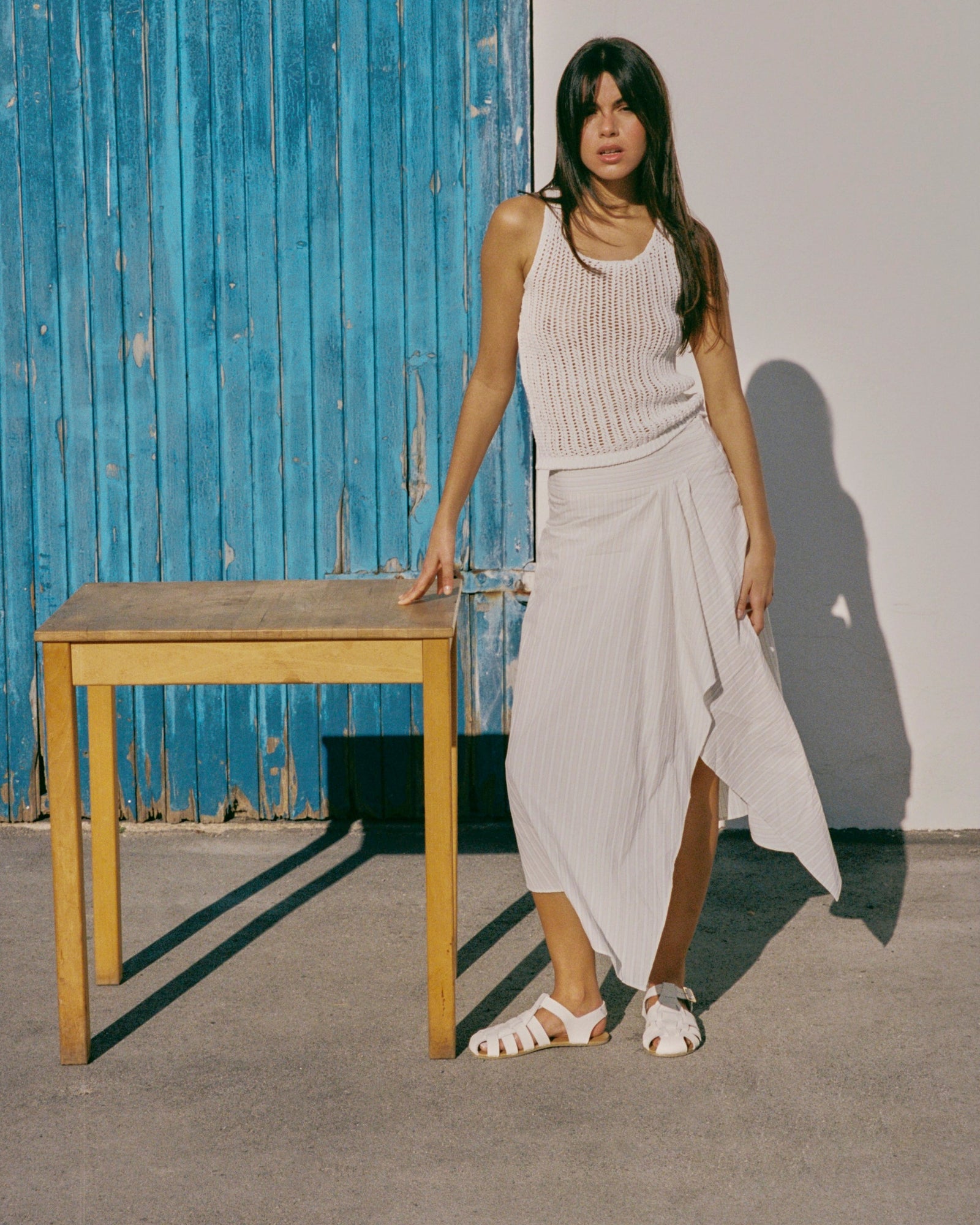 A woman stands outdoors next to a wooden table, wearing a white sleeveless ribbed knit top and a matching white wrap-style midi skirt with white sandals. The background features a blue corrugated metal door and a white wall.